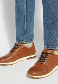 Brown leather sneakers with perforated detailing, lace-up front, white midsole, and textured accents. Worn with blue jeans and socks.