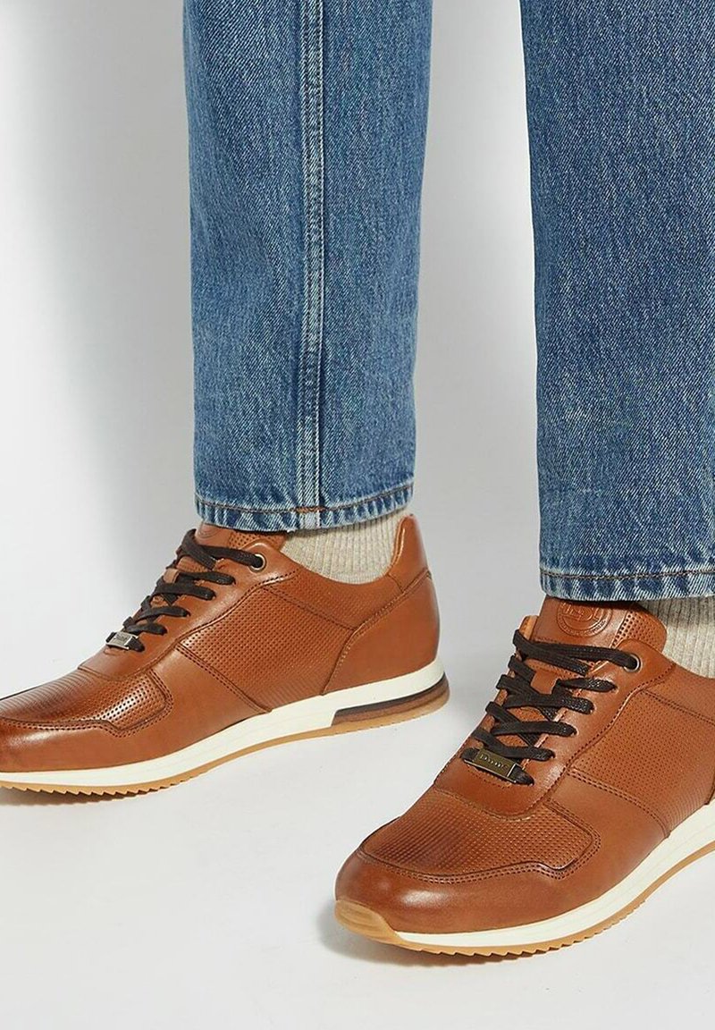 Brown leather sneakers with perforated detailing, lace-up front, white midsole, and textured accents. Worn with blue jeans and socks.