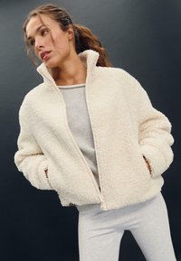 Krema barve flis jakna na zadrgo z visokim ovratnikom in puhasto teksturo, v kombinaciji z svetlo sivo majico in usklajenimi sivimi hlačami.