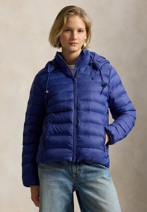 Jeune femme portant une doudoune bleue à capuche et un jean bleu clair, debout avec une main dans la poche devant un fond uni.