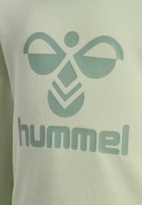 Jasnozielona bluza z podniesionym logo w ciemniejszym odcieniu zieleni, wykonana z teksturowanej tkaniny i z prążkowanym dekoltem. Projekt logo zawiera abstrakcyjne kształty.