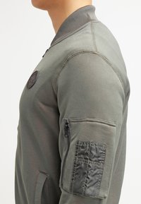 Giacca bomber verde con colletto a coste, polsini elasticizzati e tasca con zip sul braccio sinistro. Tessuto morbido e opaco con cuciture a contrasto.