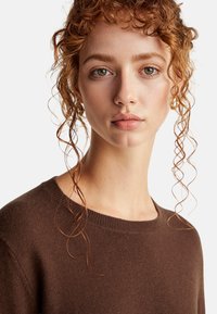 Brązowy sweter z dzianiny o okrągłym dekolcie, miękkiej fakturze i prążkowanych detalach. Modelka ma kręcone, luźne włosy opadające na twarz.