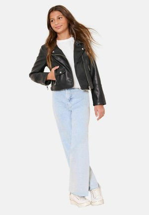 Dospievajúca dievčina s dlhými vlasmi, oblečená v čiernej koženej bunde, bielom tričku, svetlomodrých džínsach a bielych teniskách, stojaca s rukou v kapse.