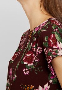 Top floreale borgogna con maniche corte, caratterizzato da motivi di fiori rosa e verdi, scollo rotondo e una texture di tessuto leggero.