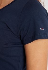 Mörkblå bomulls-T-shirt med V-ringning, kortärmar och en liten broderad logotyp med röda och vita accenter på ärmen.