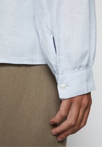 Lyseblå linned skjorte med en manchet, der har en enkelt hvid knap. Glat tekstur og afslappet pasform, parret med beige stof nedenfor.