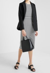 Kobieta ubrana w czarną marynarkę na białą i czarną w paski sukienkę midi, trzymająca czarną torebkę i nosząca czarne sandały na obcasie.