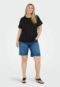 Camiseta de algodón negra de manga corta, con encaje en los bordes. Combinada con pantalones cortos de mezclilla azul, que terminan justo por encima de la rodilla, y sandalias negras de tacón.