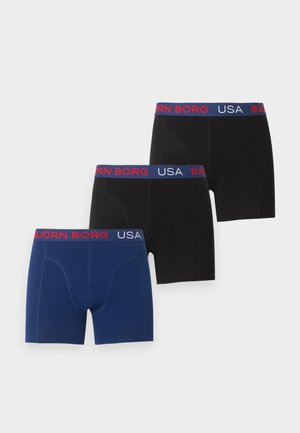 Manlijk model staat op blote voeten, draagt zwarte boxershorts met een blauw-rode tailleband met het opschrift "BJORN BORG USA", op een effen witte achtergrond.