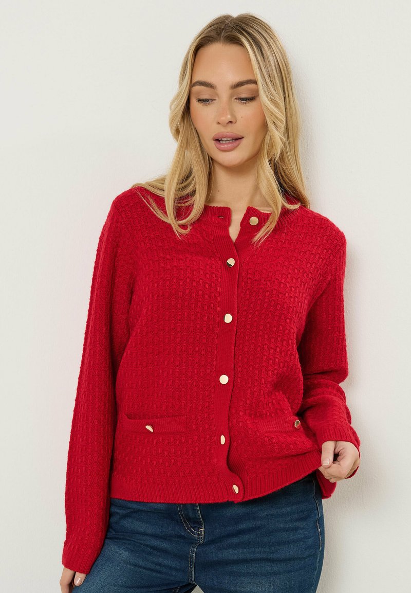 Mujer con un cárdigan rojo texturizado abotonado con botones dorados y bolsillos frontales, combinado con vaqueros azul oscuro, posando frente a un fondo blanco.