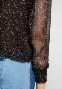 Blusa nera trasparente con motivo di stelle colorate in rosa, arancione e bianco. Presenta polsini arricciati e una vestibilità morbida. Sfondo in denim visibile.