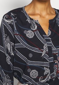 Blusa blu navy con motivi paisley e geometrici in bianco, rosso e blu. Presenta una chiusura frontale e maniche leggermente a sbuffo.