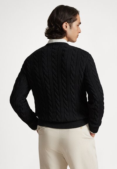 Άνδρας με σκούρα κυματιστά μαλλιά, ντυμένος με μαύρο πουλόβερ ριπ με πλεκτό σχέδιο και ανοιχτόχρωμο παντελόνι, στέκεται με τα χέρια στις τσέπες, στραμμένος πλάτη.