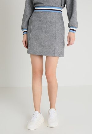 Femme portant une mini-jupe grise avec une ceinture rayée bleue, un haut gris à manches longues assorti, et des baskets blanches, debout sur un sol blanc.