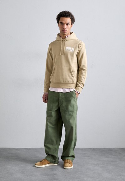 Sweatshirt à capuche beige avec poche avant et logo "POLO", porté par-dessus une chemise rose clair. Un pantalon ample vert olive et des baskets beiges complètent le look.