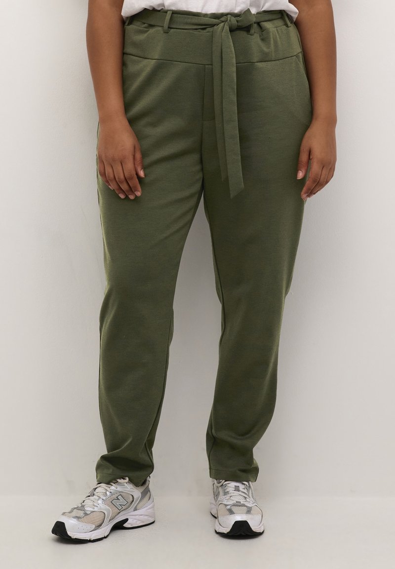 Personne portant un pantalon vert olive à la taille nouée et des chaussures de sport blanches, debout devant un fond neutre.