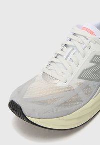 Baskets de sport gris clair avec une tige en mesh texturé, des accents réfléchissants et une semelle extérieure en caoutchouc noir. Elles présentent des lacets fins blancs et une semelle rembourrée.
