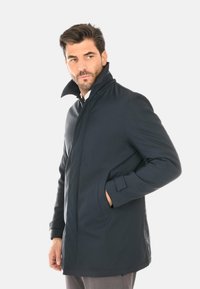 Cappotto navy con colletto alto, maniche lunghe e tasche laterali. Realizzato in un materiale liscio e resistente alle intemperie, presenta un design minimalista.