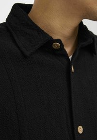 Camisa negra texturizada con cuello, que presenta botones de madera y un patrón de espiga visible en la tela.