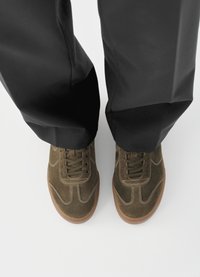 Αθλητικά παπούτσια από σουέτ σε ελιά πράσινο με ανοιχτό ράψιμο, καφέ ελαστική σόλα και σχέδιο με κορδόνια, συνδυασμένα με μαύρο παντελόνι, από ψηλά.