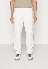 Pantalones de chándal blancos con franjas laterales verdes, cinturilla elástica y diseño de pierna ajustada, fabricados en tejido liviano. Combinados con zapatillas blancas.