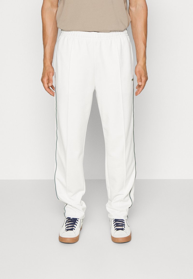 Pantalones de chándal blancos con franjas laterales verdes, cinturilla elástica y diseño de pierna ajustada, fabricados en tejido liviano. Combinados con zapatillas blancas.