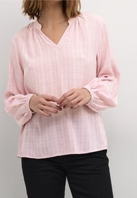 Blouse rose clair avec un col en V, tissu texturé présentant des motifs discrets à carreaux, et manches longues froncées. Associée à un pantalon noir.