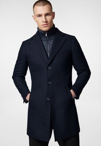 Granatowy płaszcz wełniano-syntetyczny o wyraźnej sylwetce, z klapami w szpic i zapięciem na trzy guziki. Posiada kieszenie boczne oraz kontrastowy, prążkowany wewnętrzny kołnierz.
