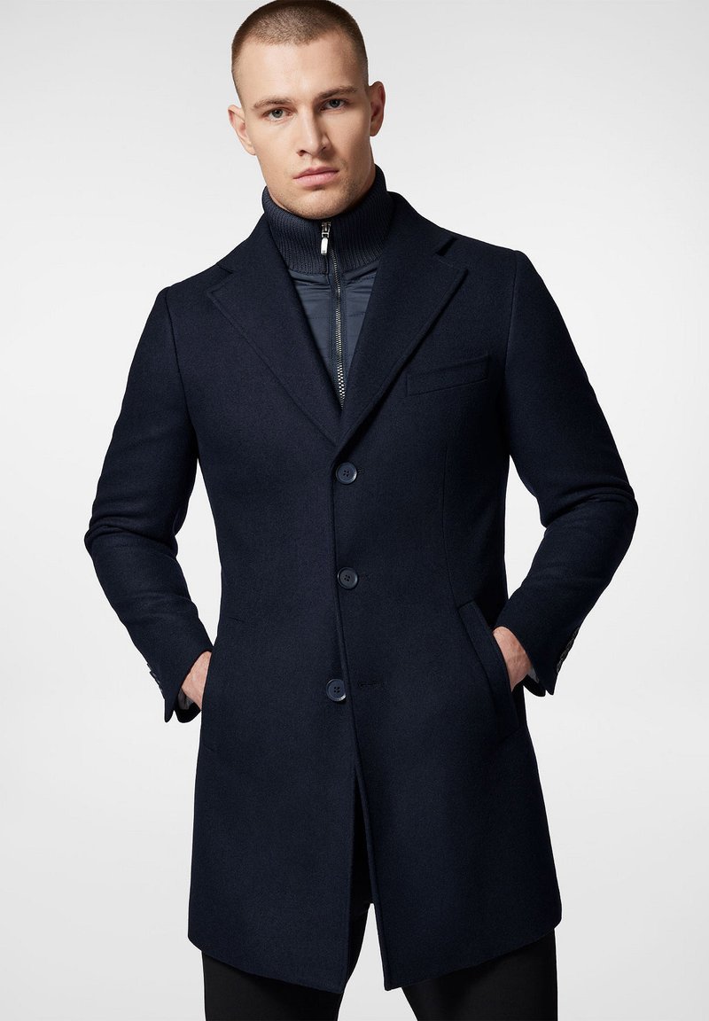 Granatowy płaszcz wełniano-syntetyczny o wyraźnej sylwetce, z klapami w szpic i zapięciem na trzy guziki. Posiada kieszenie boczne oraz kontrastowy, prążkowany wewnętrzny kołnierz.