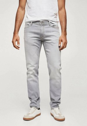 Homme portant un jean slim gris clair, des baskets blanches à semelles marron, et un t-shirt blanc rentré, debout devant un fond uni.