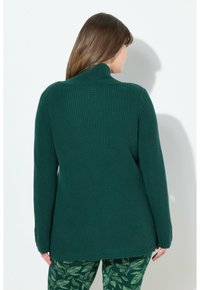 Grüner Strickpullover mit stehem Kragen, gerippter Textur und langen Ärmeln, kombiniert mit gemusterten grünen Leggings mit Blättern.