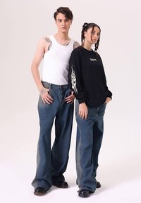 Dva modelové v ležérních outfitech: jeden v bílém tílku a modrých džínách, druhý v černé mikině s plamenovými potisky a modrých džínách.