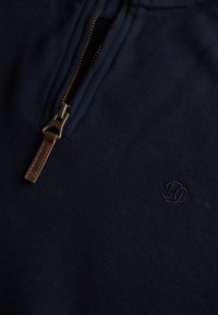Marineblå bomulds-pullover med en kvart lynlås. Har en metal lynlås og en læderdetalje på trækknappen. Broderet logo på brystet.