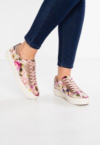 Blommönstrade sneakers med rosa satinfinish, kontrasterade med roséguldsaccent och vita sulor, sedd från sidan mot en neutral bakgrund.
