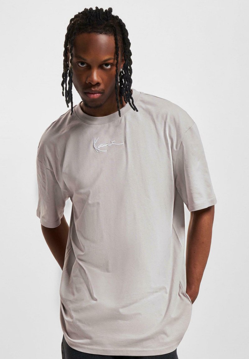 T-shirt gris à manches courtes avec col rond, featuring broderie cursive blanche sur la poitrine et une coupe décontractée. Texture de tissu lisse.