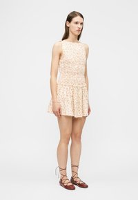 Γυναίκα που στέκεται φορώντας ένα αμάνικο, ανοιχτό ροζ φόρεμα με μικρές κόκκινες βούλες και καφέ παπούτσια με κορδόνια, μπροστά σε ένα απλό ανοιχτό φόντο.