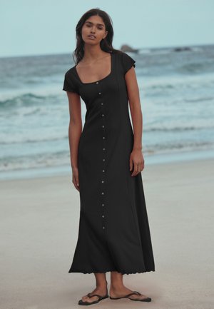 Nő áll a tengerparton, fekete rövid ujjú, gombos maxi ruhát és fekete papucsot viselve, mögötte az óceán hullámai.