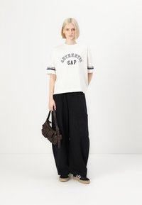 Bílé oversized tričko s potiskem "AUTHENTIC GAP", v kombinaci s černými širokými cargo kalhotami. Hnědá taška přes rameno a černé tenisky s bílými pruhy.