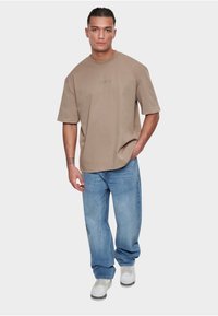 Tan short-sleeve t-shirt with a relaxed fit, paired with loose blue jeans and white sneakers. Subtle logo detail on the shirt's chest.