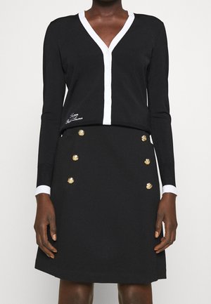 Vrouw draagt een zwart vest met lange mouwen, witte bies en geborduurde tekst, gecombineerd met een zwarte rok met twee rijen gouden knopen.