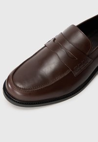 Braune Leder-Slipper mit glatter Textur, ausgestattet mit einem dekorativen Riemen und einer Naht auf dem Vorderblatt sowie einer abgerundeten Zehenpartie.