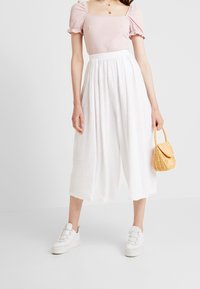 Vita vida culotter med texturerad yta, kombinerat med en ljusrosa åtsittande topp. Accentuerad med en liten vävd gul handväska och vita sneakers.