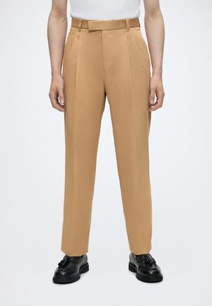 Beige skræddersyede bukser med folder, båret med sorte mokkasiner med kvaster og en hvid skjorte, vist på en stående person fra taljen og ned.