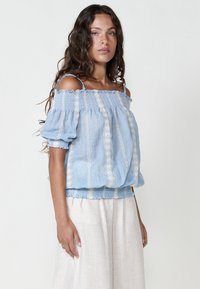 Blusa azzurro chiaro, senza spalle, con dettagli smock, accenti ricamati e maniche a sbuffo, abbinata a pantaloni crema larghi.