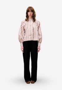 Vrouw staat rechtop met het gezicht naar voren, gekleed in een gestreepte blouse met knoopsluiting en puffmouwen, zwarte broeken en bruine schoenen, op een witte achtergrond.