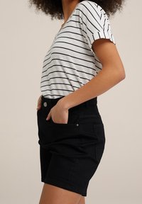 Frau in einem weiß-schwarz gestreiften kurzärmeligen Oberteil, das in schwarze hochgeschnittene Jeansshorts gesteckt ist, Hand in der Tasche, beiger Hintergrund.