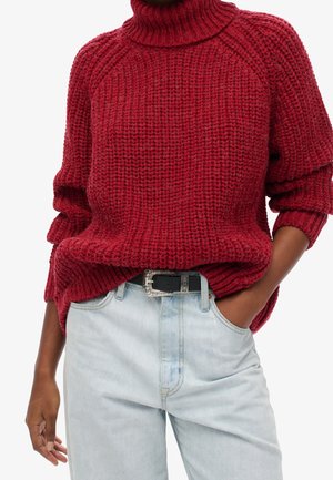 Personne portant un pull à col roulé rouge épais, rentré dans un jean bleu clair avec une ceinture noire décorative, main dans la poche.