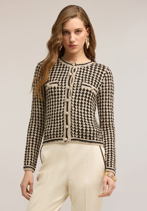 Kobieta ubrana w czarny i beżowy sweter na drutach w wzory, zapinany na perłowe guziki, kremowe spodnie z wysokim stanem, złote kolczyki w kształcie liści oraz bransoletki.