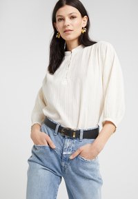 Blusa de crema clara con rayas verticales texturizadas, un escote abotonado y mangas abullonadas, combinada con jeans azul claros de cintura alta y un cinturón negro.